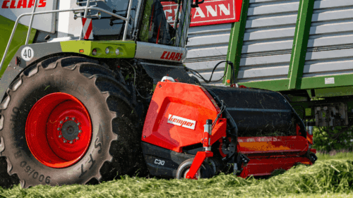 A green and white Claas forage harvester unloading into a large green and grey trailer. The machine has a red Kemper grass collecter on the front.