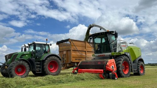 A red Kemper grass pick up attachment connected to a Claas forage harvester, collecting grass in a field next to an orange Richard Wester trailer. The trailer is being pulled by a Fendt tractor whilst silage is unloaded into the trailer behind.